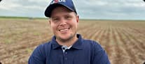 João Pierobon usando camisa polo azul e boné em área rural com campo ao fundo.