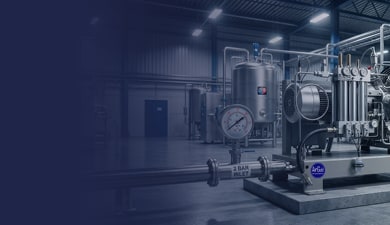 Compressor industrial em fábrica com tubulações, manômetros e equipamentos metálicos ao fundo.