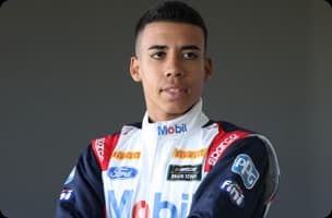 Murilo Rocha usando camisa azul e boné da Mobil em frente a prédio com detalhes vermelhos.