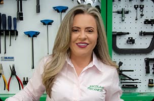 Sandra Nalli, fundadora da Escola do Mecânico, usando camisa branca em ambiente de oficina com equipamentos verdes ao fundo.