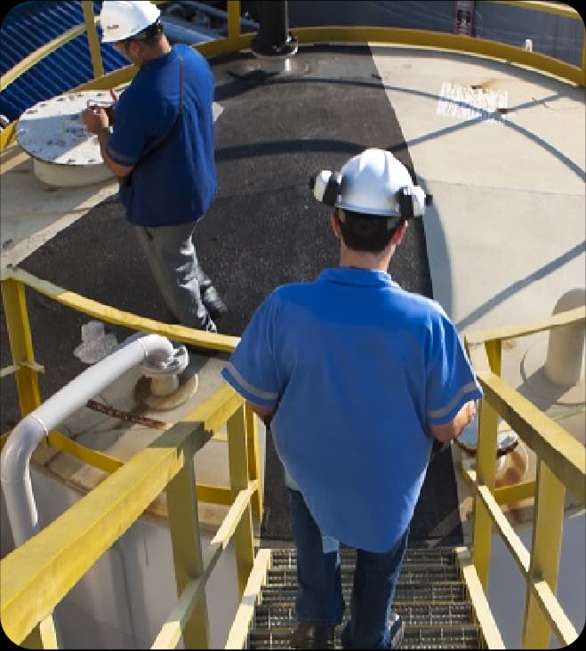 Dois profissionais usando capacete e uniforme azul em estrutura de tanque industrial com guarda-corpo amarelo realizando inspeção em vista de cima.