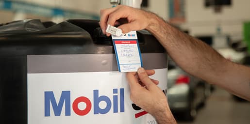 Mãos aplicando etiqueta de identificação em tanque de óleo Mobil dentro de oficina automotiva.